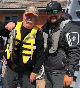 Bruce Roulston posing with a caught fish on a dock beside Eugene Chong at CNIB Lake Joe. They are both wearing life jackets and a ballcap.
