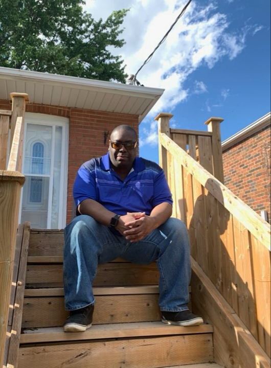 Martin sits on the porch steps outside his home. He’s wearing a blue polo t-shirt, jeans and sunglasses.