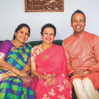 Prema, a South Asian woman with curly hair, wears a pink saree and sits next to two people – her friend Chamundi Eswari Selvaraj, a South Asian woman with long hair, wearing a green and blue plaid saree and her son Prasanna, a South Asian man with short hair, who is wearing a pink kurta. They are all seated on a green couch