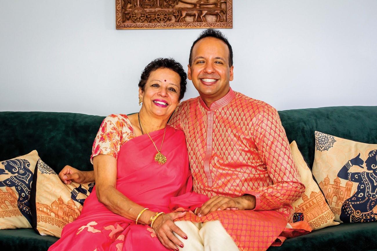Prema, a South Asian woman with curly hair, wears a pink saree and sits next to her son Prasanna, a South Asian man with short hair, who is wearing a pink kurta. They are both seated on a green couch with yellow printed throw pillows.