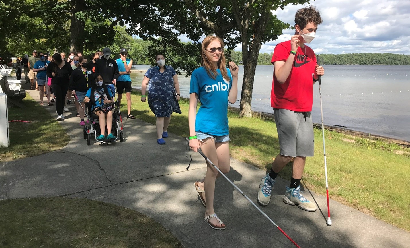 Two youth with white canes are walking along the lakeshore at CNIB Lake Joe. A group of additional youth are walking behind them.