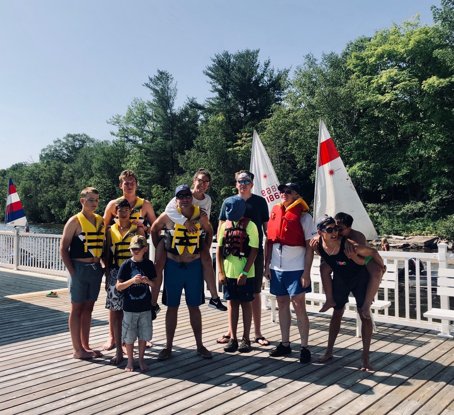 A small group of people standing on the dock in shorts and life jackets. They are smiling and, in some cases, piggy-backing others