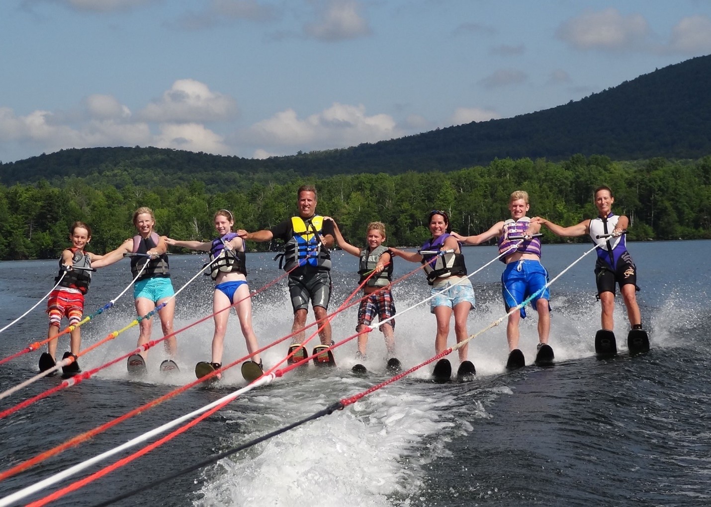 A photo of 8 people - men, women, girls and boys - waterskiing together in line with their arm on the person behind them.