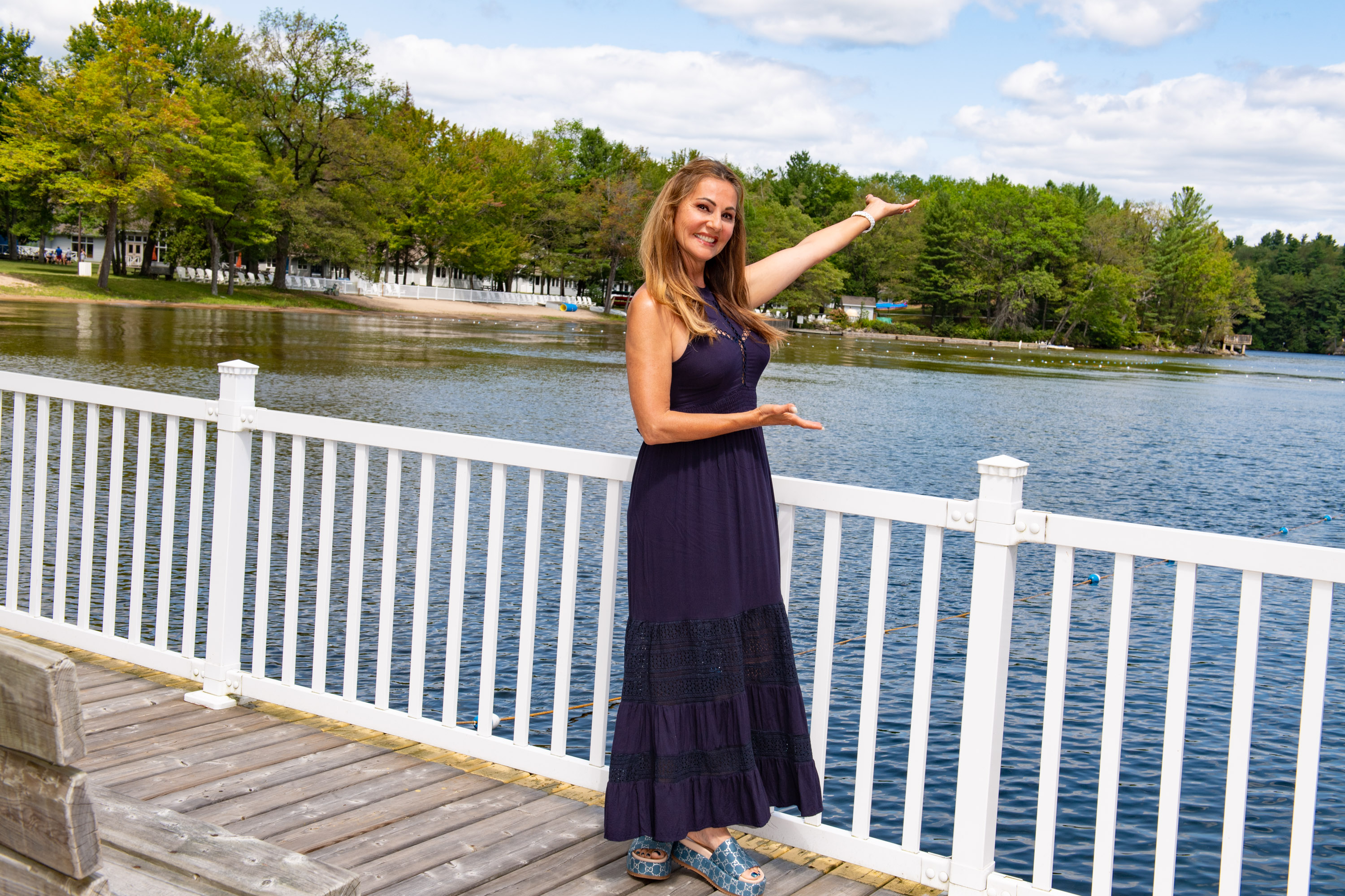 Joan Kelley Walker standing on the CNIB Lake Joe boathouse deck, smiling with her arms open and pointing toward the shoreline.