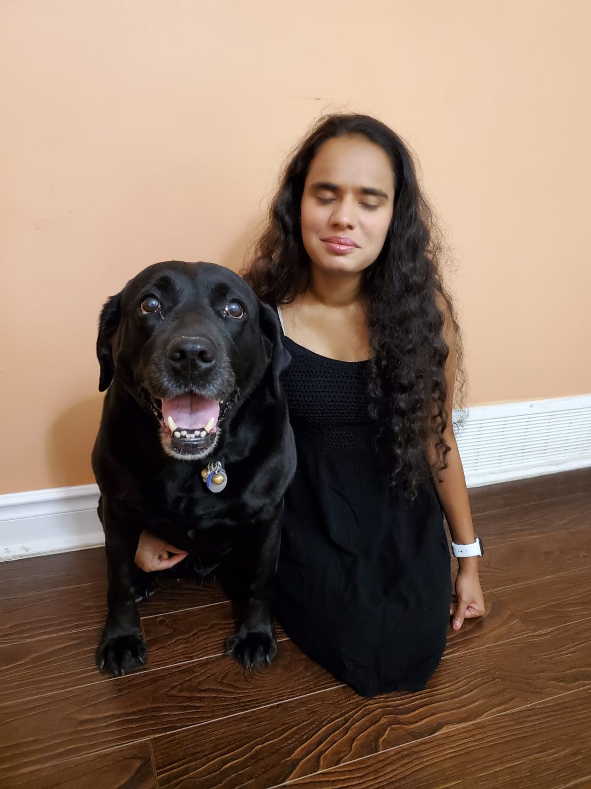 Runa Patel, kneeling on the floor next to her guide dog, smiling for the camera.