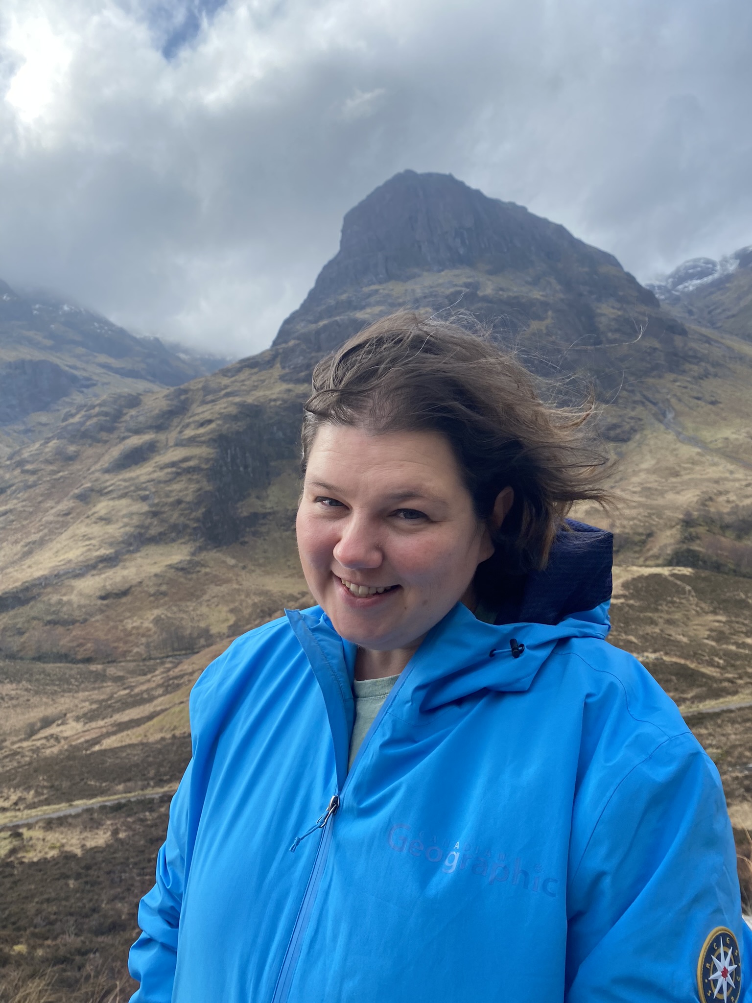 Sam Smadella is smiling standing in front of a mountain range. Sam is wearing a blue wind breaker jacket, and her hair is blowing to the side from the wind.