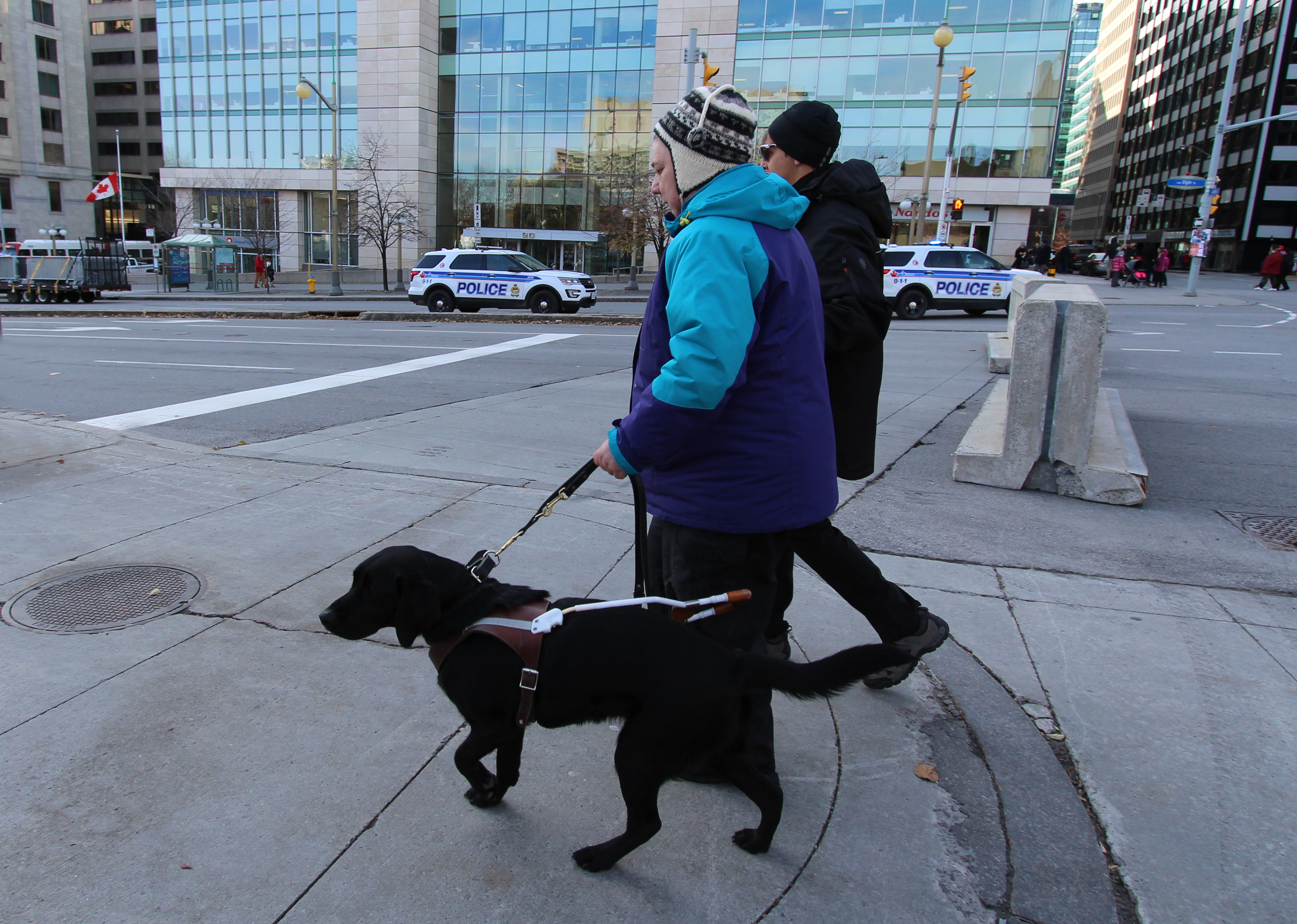 Sandy and her guide dog Keller, walking along a sidewalk in a busy downtown area during a cold day.