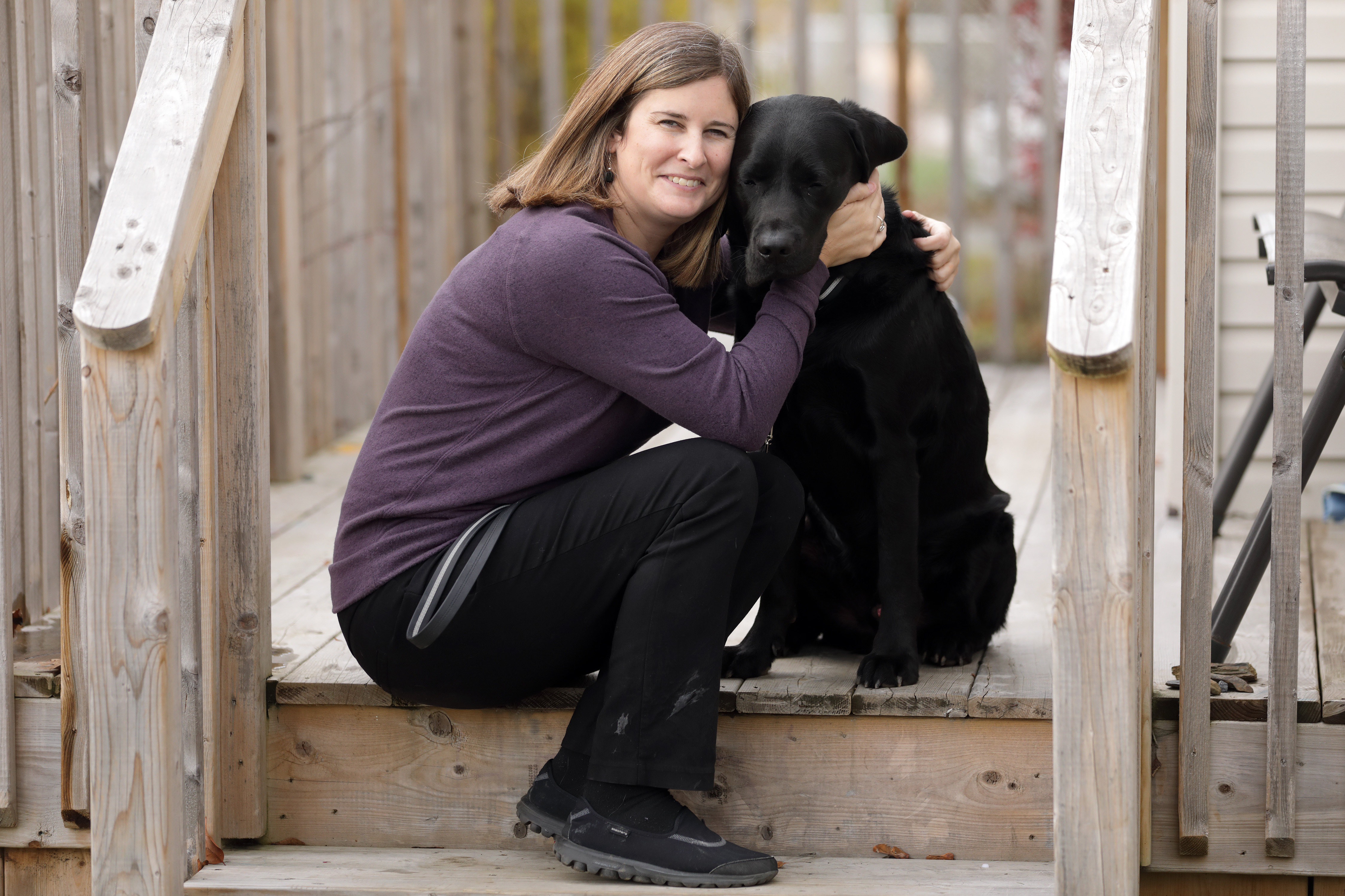 Photo of Shelley sitting on a wooden porch, smiling while giving Rookie a big hug. 