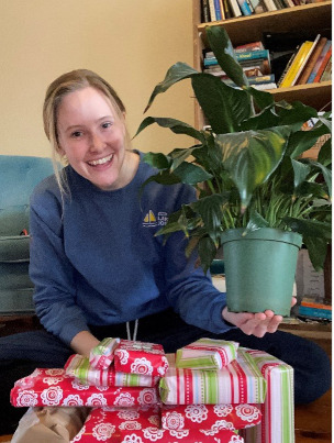 Amy is kneeling in front a pile of wrapped presents, holding one of her houseplants overtop while smiling.