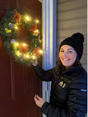 Lindsay is smiling at her front door, pointing to her holiday wreath that is brightly lit.
