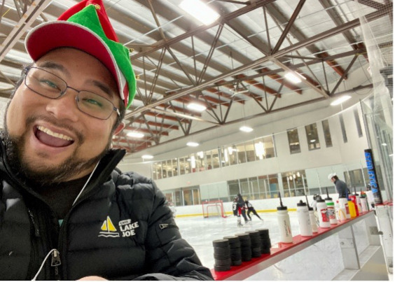 Eugene is at a local hockey arena wearing a red & green elf hat on top of his baseball hat.]