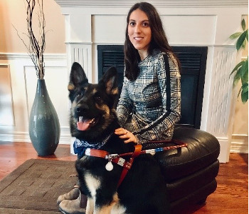 Stela sitting on a leather ottoman in front of a fireplace with her German Shepherd guide dog in front of her. 