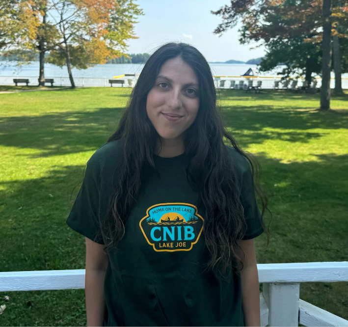 Stela se tient sur la promenade du camp Lake Joe d'INCA et elle tourne le dos au lac. Elle porte un t-shirt vert olive du camp Lake Joe d'INCA. 