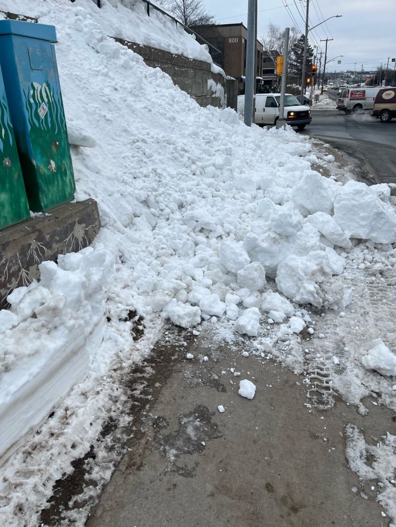 Un banc de neige bloque une partie du trottoir, obligeant les piétons à marcher sur la route. Une portion de trottoir dégagée qui disparaît sous un gros tas de neige d'environ 3 pieds sur la gauche et de 2 pieds sur la droite. 