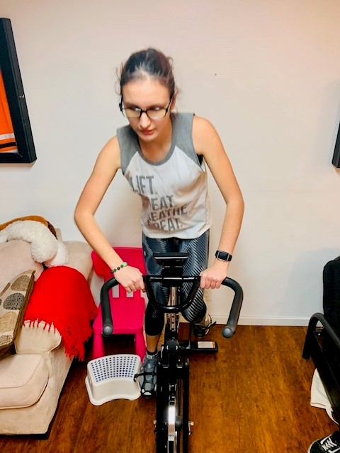 Veda, wearing athletic clothes and glasses, rides her indoor spin bicycle. 
