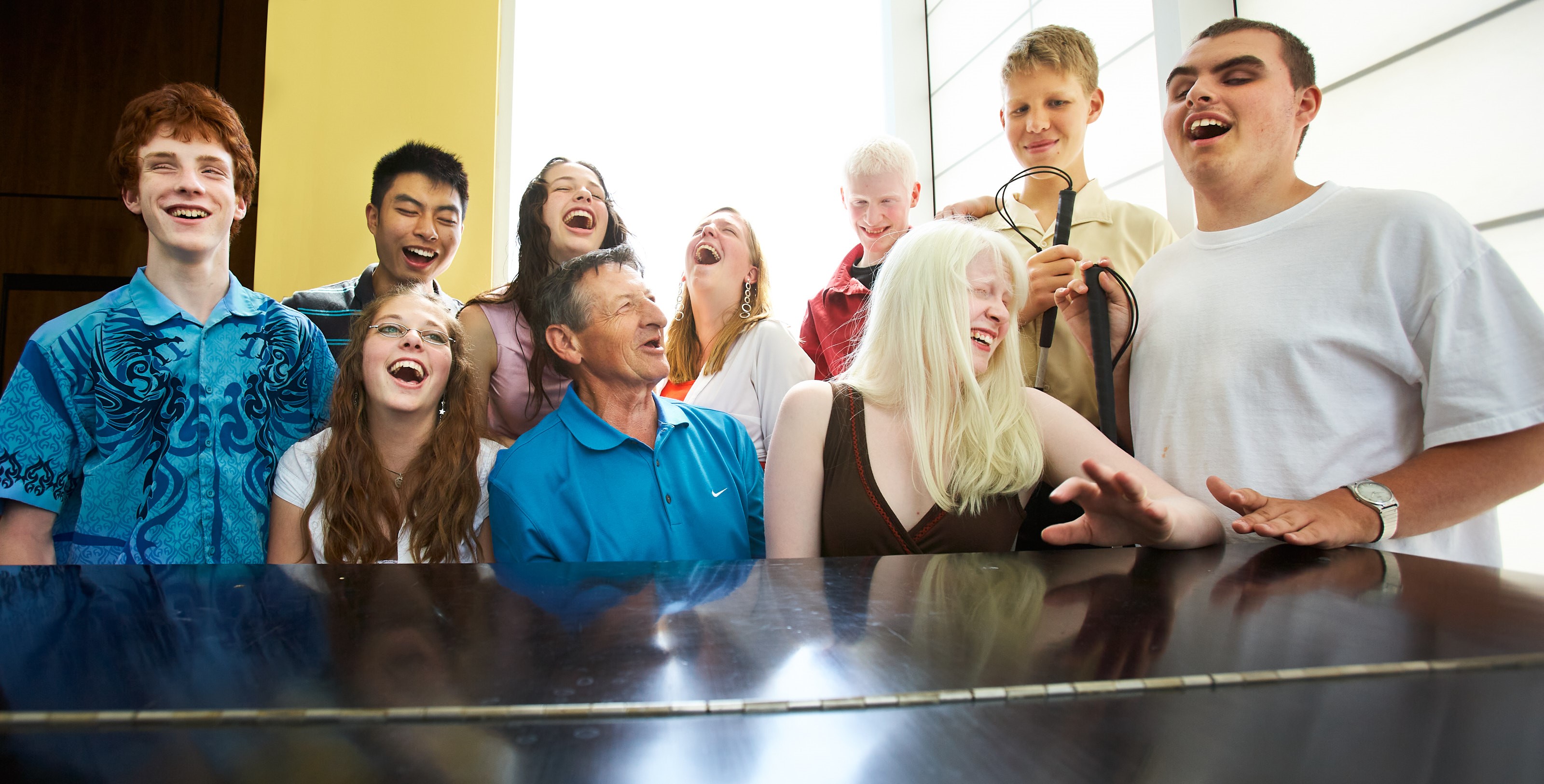 Walter Gretzky sits at a grand piano surrounded by 8 youth with sight loss. They are smiling and excitedly singing a tune while Walter plays the piano.