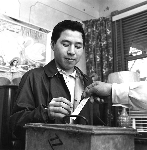 Une photo en noir et blanc d'un homme recevant son bulletin de vote avec une boîte de scrutin métallique au premier plan.  