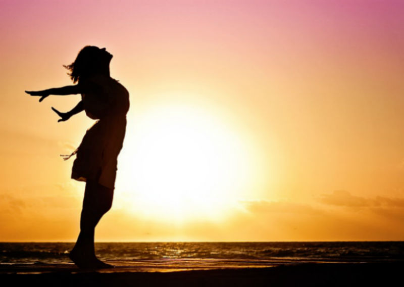 A woman stands silhouetted against a sunset, arms outstretched 