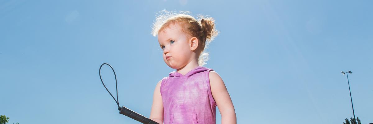 Little girl stands on tactile marker holding her white cane. She's wearing a purple dress and matching shoes.