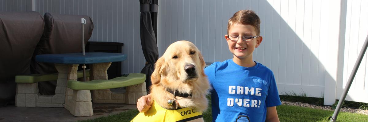A smiling boy in glasses with his arm around a Golden Retriever in a yellow vest.