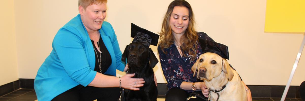Ashley and Danika with two Lab/Golden Retriever crosses (one black and one yellow) wearing graduation caps.
