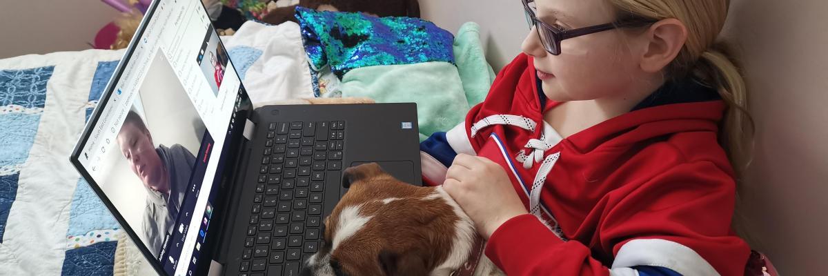 Madelyn sits in bed with her small puppy and laptop on her lap. She is participating in a Zoom call. 