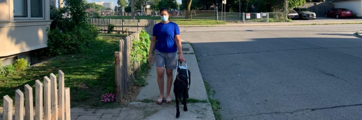 Victoria Nolan and her guide dog, Alan, walking along a sidewalk on a sunny day. Victoria is wearing a face mask.
