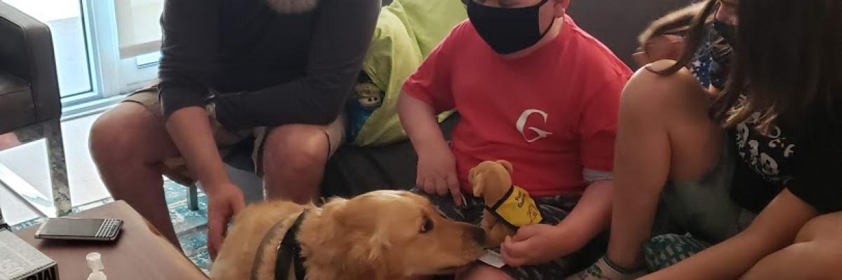Ollie and family members sitting on a couch wearing face masks. Ambassador Dog Ziggy is sitting on the floor in front of Ollie, sniffing the stuffed CNIB Guide Dog plush puppy on Ollie's lap.