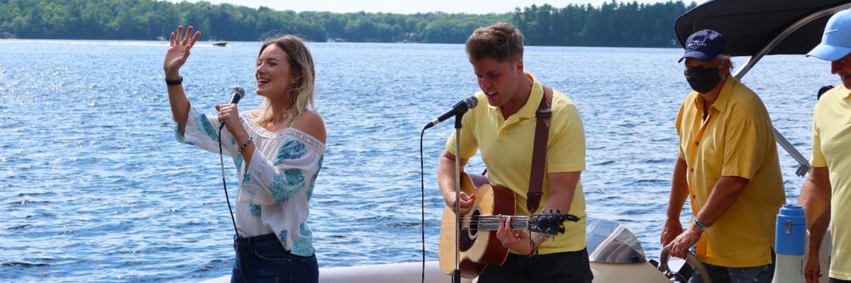 Leah Daniels singing and waving with Will Hebbes beside her playing guitar on a party pontoon, entertaining docks guests on Lake Muskoka.