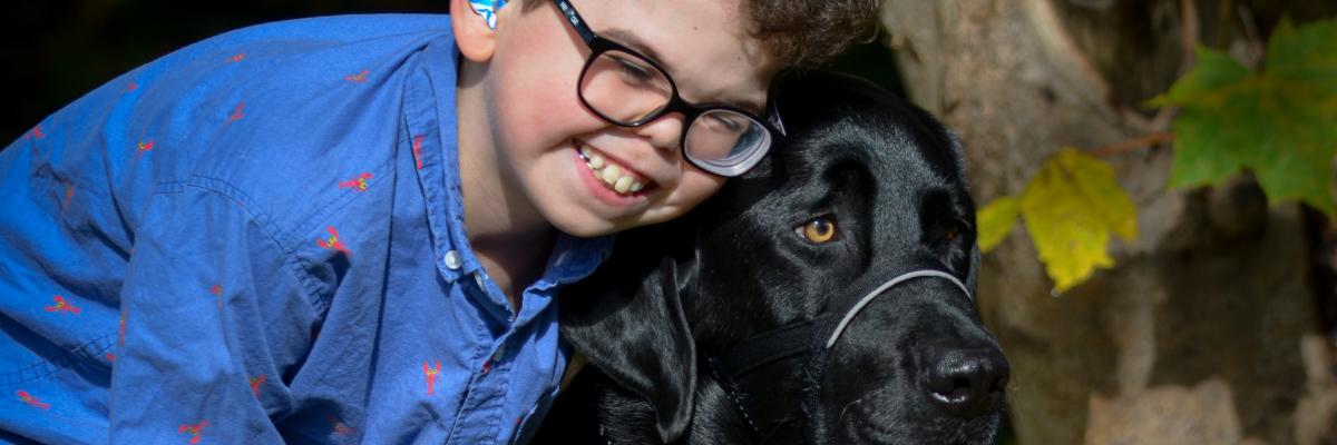 10-year-old Kaiden smiling and leaning down to embrace his CNIB Buddy Dog, Flinn, a black Labrador-Retriever cross.