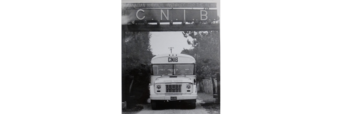A black and white photograph of an old school bus with "CNIB" on the front, driving under the "CNIB" welcome signage at Lake Joe.