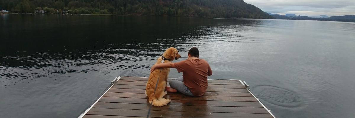 Landon and Ruggles, a golden retriever, sitting on a wharf overlooking a lake; Landon’s arm is around Ruggles who is looking at Landon.