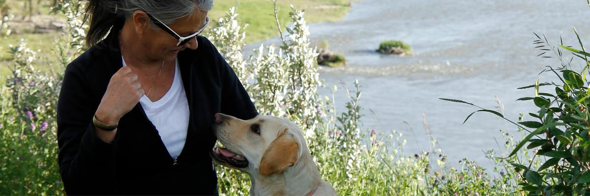 Debra Williamson kneeling next to Patsy, a yellow Labrador-Retriever wearing a Future Guide Dog vest, exchanging a glance with the other in front of the Bow River in Calgary