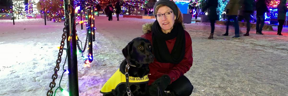 Lorraine Rempel kneeling on the snowy ground next to Jennie, a black Labrador-Retriever cross wearing a yellow Future Guide Dog vest. Trees decorated in lights are behind them.