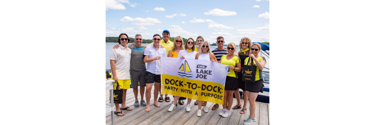 CNIB Muskoka Dock-to-Dock event volunteers posing at the CNIB Lake Joe boathouse, holding event flags.