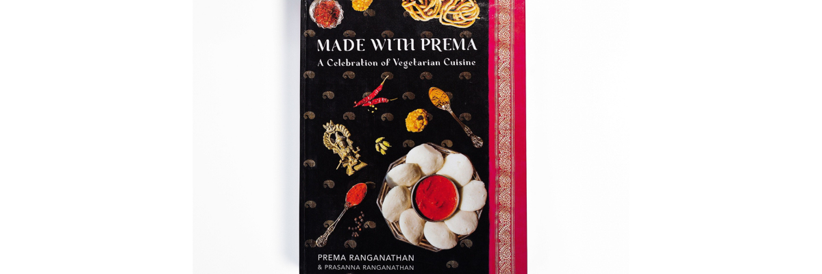 Une photo d’un livre de cuisine à couverture rigide intitulé  Made with Prema apparaît sur un fond blanc uni. La couverture du livre de cuisine présente plusieurs plats sur un sari noir, or et magenta