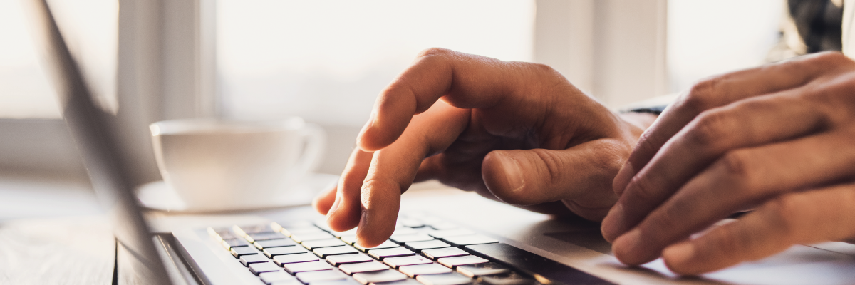 Gros plan sur les mains d'une personne tapant sur le clavier d'un ordinateur portable. L'accent est mis sur les mains et le clavier, l'arrière-plan est flou mais il s'agit d'un intérieur bien éclairé