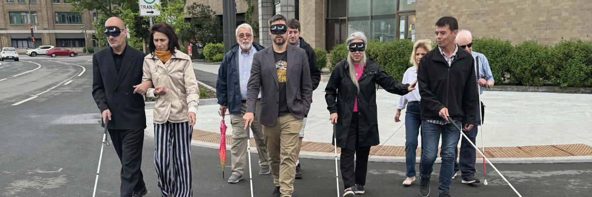 Claude les yeux bandés guidé par Valérie directrice d’INCA Québec, Pierre-Luc les yeux bandé guidé par Jean-Michel et un bénévole derrière lui, Maude guidée par David et René du RPHV 03-12 à l’arrière traversent la rue en direction de la caméra en balayant leur canne-blanche devant eux