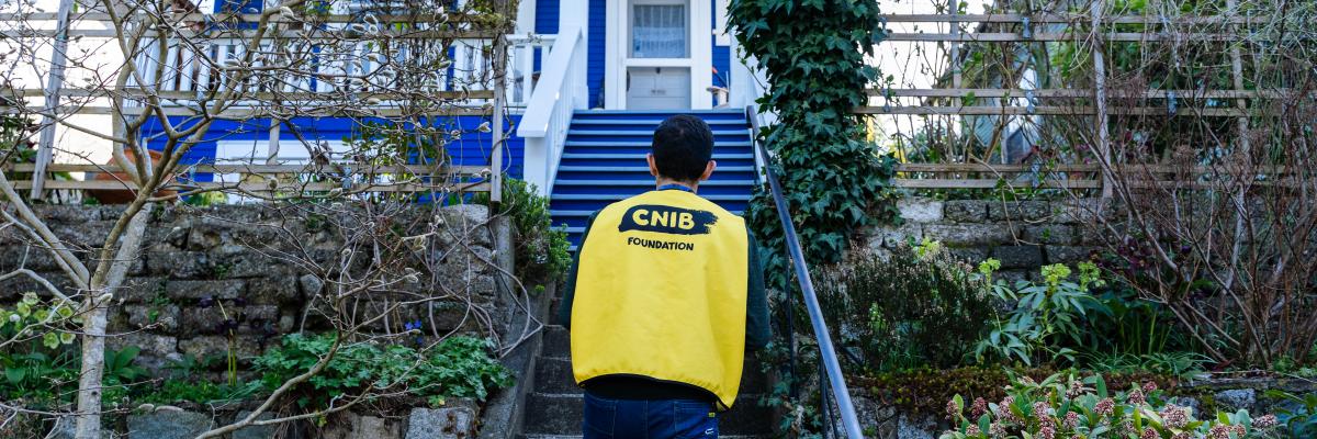 Un collecteur de fonds portant un gilet jaune muni du logo de la Fondation INCA au dos monte un escalier, au milieu d'un jardin verdoyant, menant à la porte d'entrée d'une maison.