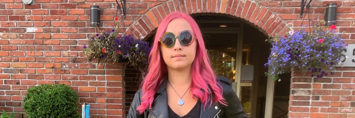 A cool teenager stands outside her house with her white cane in hand. She has pink hair and is wearing a leather jacket.