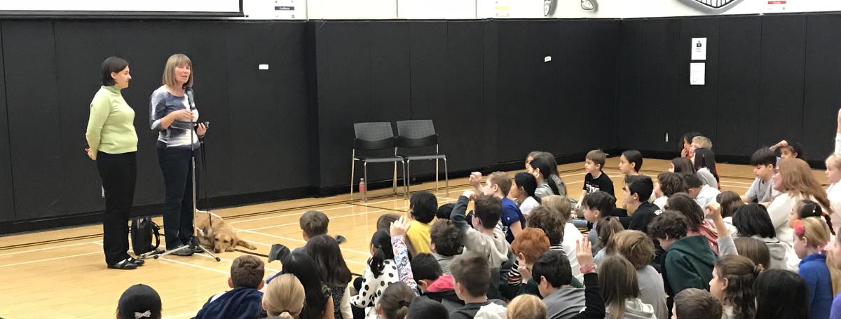 An ambassador stands at the front of a room and gives a presentation to 30+ seated children.