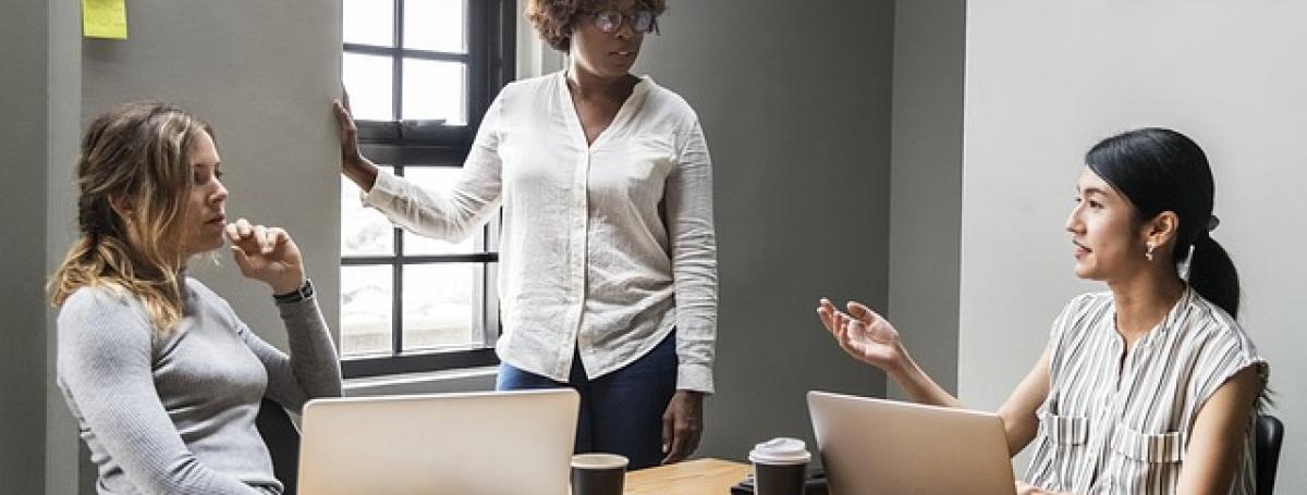 Three women in a meeting. Two women are sitting at a table in front of a laptop. Another woman stands near a window. They are engaging in a serious conversation.