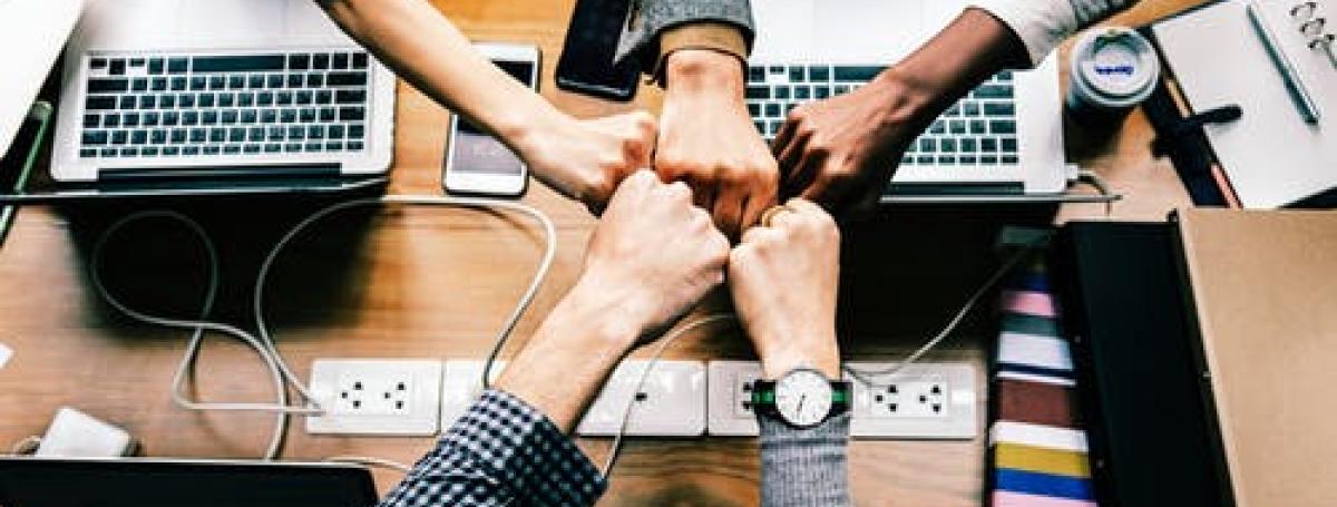 5 people but their fists together in the middle of a table covered in computers, cords, and phones.