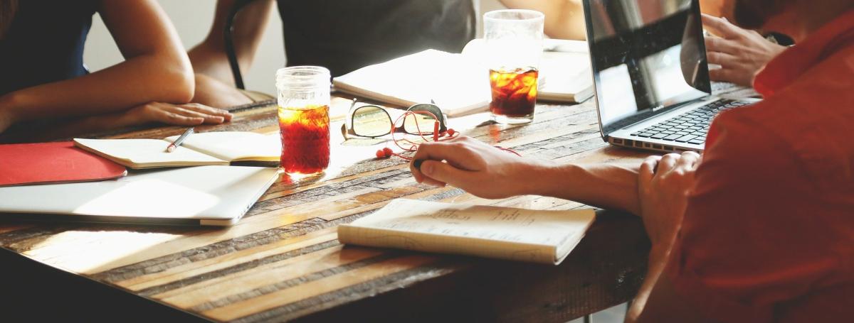an extreme close up of 4 people sitting at a wooden table. A laptop, paper, pens, glasses, notebooks are infront of them