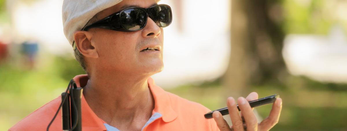 A man wearing sunglasses and a hat holds an iPhone in his left hand. A white cane sits in his right hand.