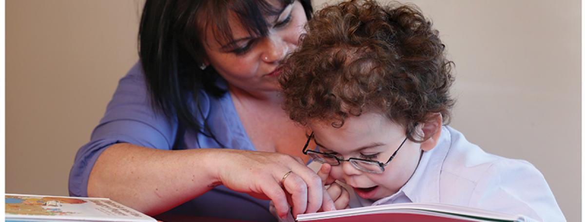 A young boy reads a tactile book. His mother helps guide his hands over the book.