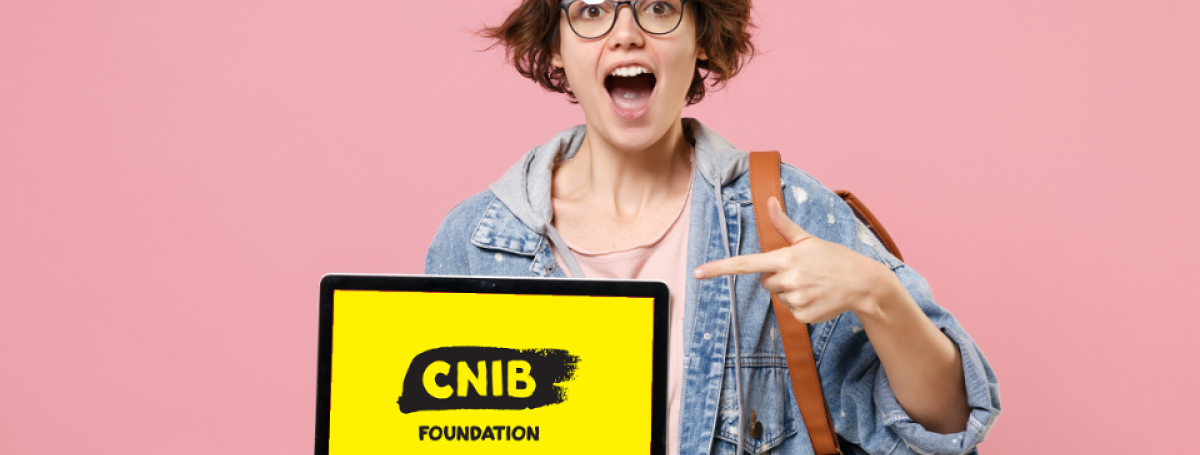 A young teenage girl excitedly holds a laptop monitor displaying the CNIB Foundation logo. She is pointing to the screen with her left hand.