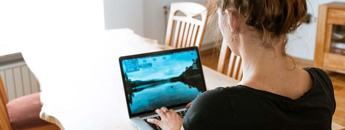 The back of a young woman's head. She sits at kitchen table and types on her laptop computer.