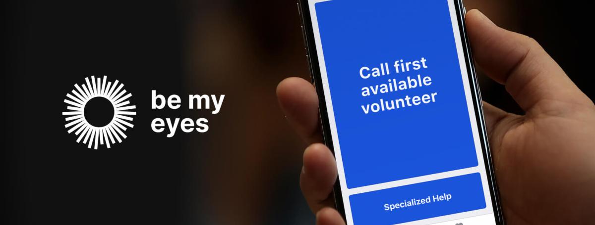 A hand holds a smartphone up in the air. The "Be my Eyes" app is open on the homescreen.