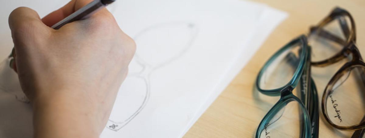 Photo of a person's hand, sketching on a white piece of paper with two pairs of glasses on the right.